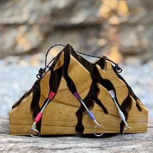 May include: Wooden mountain-shaped decor with three colorful climbing carabiners. The mountain is made of light brown wood with dark brown accents. The carabiners have red, purple, and white accents and silver key rings.