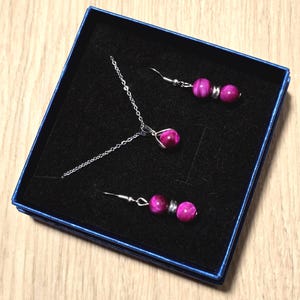 May include: A jewellery set comprising a necklace and earrings, displayed in a blue box with a black interior. The set includes a silver chain necklace with a pendant and matching earrings, all featuring vibrant pink beads.