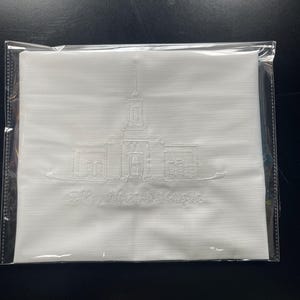 Op de afbeelding: Witte stoffen servet met een geborduurd beeld van een gebouw en de tekst "Elko Nevada Temple". Het servet is verpakt in een doorzichtige plastic hoes. Het gebouw heeft een torenspits en meerdere ramen.