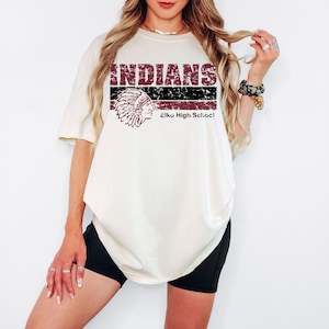 May include: An off-white short-sleeve t-shirt featuring a distressed graphic with the text INDIANS and Elko High School. The design includes a maroon and black horizontal striped pattern and a detailed illustration of a Native American headdress.