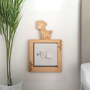 May include: A wooden light switch cover featuring a giraffe design. The light brown frame surrounds a white switch. A small giraffe figure is placed on top. The frame has leaf details. The switch is mounted on a white wall.