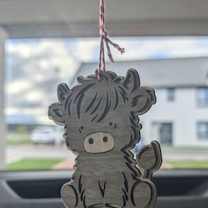 May include: A wooden Highland cow ornament, suspended by a red and white string. The cow is light brown with darker brown detailing. The ornament is hanging in front of a window, with a house visible in the background.