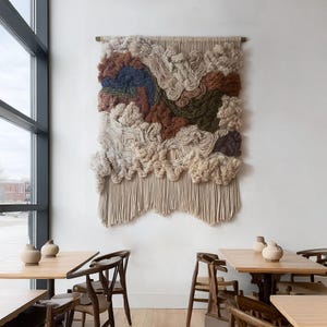 May include: A large, textured wall hanging with a neutral color palette. The artwork features a mix of beige, brown, and blue wool fibers, creating a layered, cloud-like design. The bottom edge has a fringe. The piece is mounted on a wall above a wooden table.