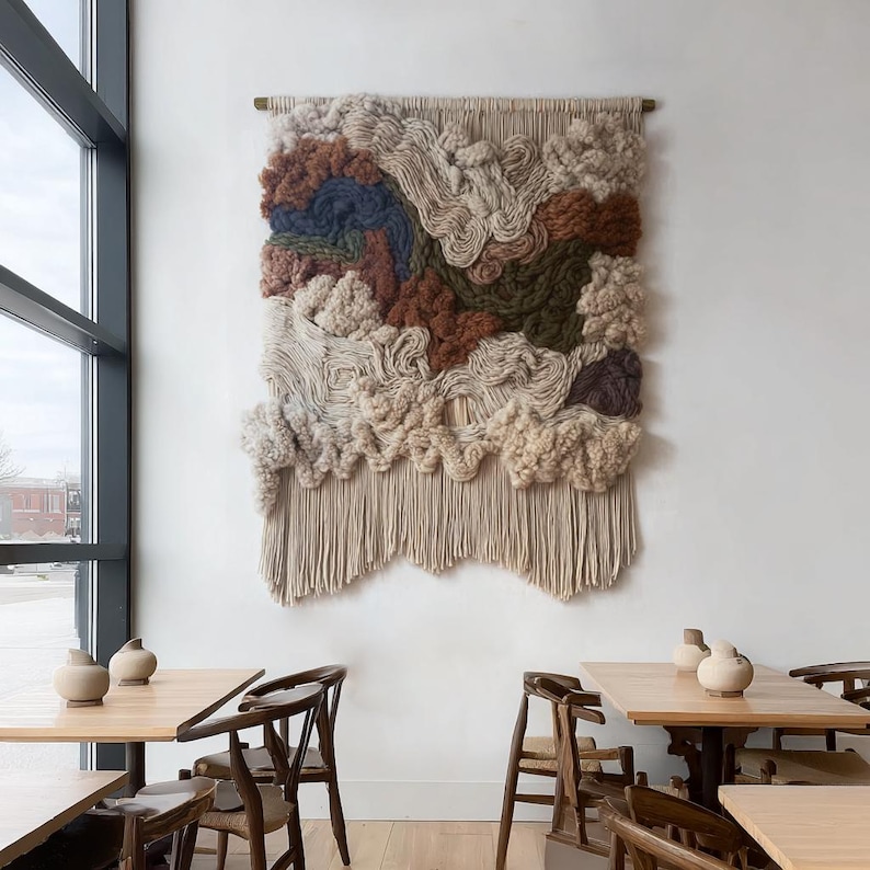 May include: A large, textured wall hanging with a neutral color palette. The artwork features a mix of beige, brown, and blue wool fibers, creating a layered, cloud-like design. The bottom edge has a fringe. The piece is mounted on a wall above a wooden table.