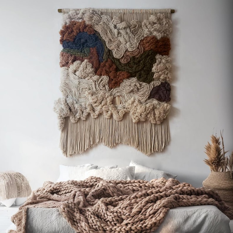 May include: A large, textured wall hanging with a neutral color palette. The artwork features various shades of beige, brown, and blue, with a fringe at the bottom. The piece is mounted on a wooden rod, creating a cozy, bohemian aesthetic.