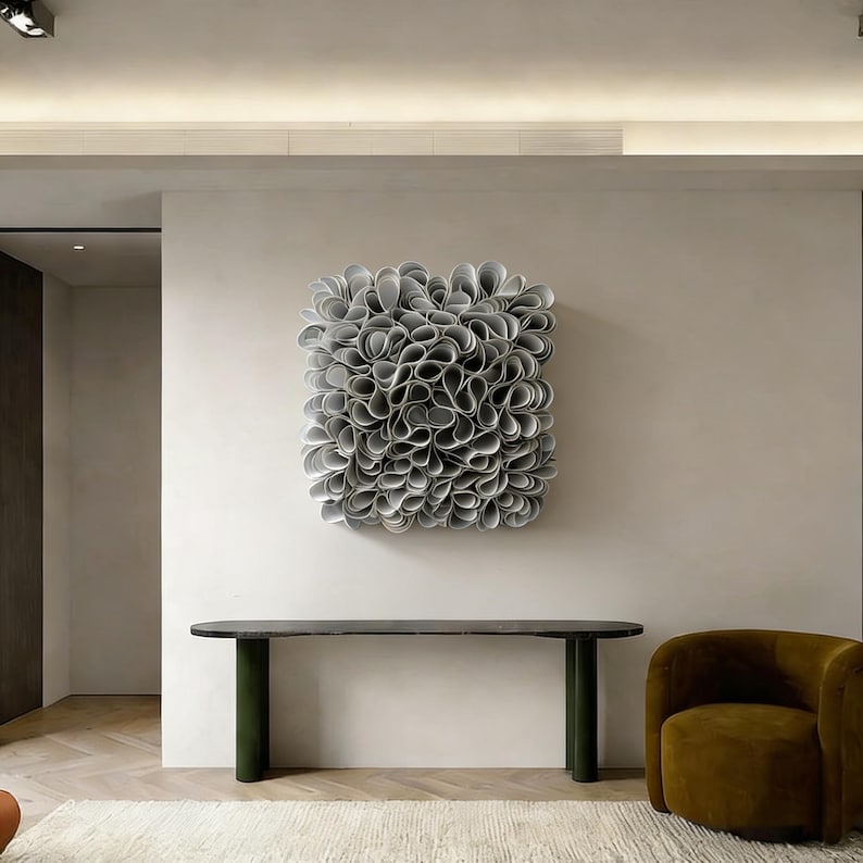 May include: Abstract wall art made of layered grey paper, mounted on a white wall. Below, a dark grey console table with green legs. A mustard-coloured armchair and cream rug complete the interior design. The artwork is approximately 100 cm x 100 cm.