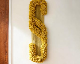 May include: Mustard yellow wall hanging with a textured, abstract design. The piece features a large, stylized 'S' shape created with a thick, looped yarn. The background is a woven, vertical panel. Home decor.