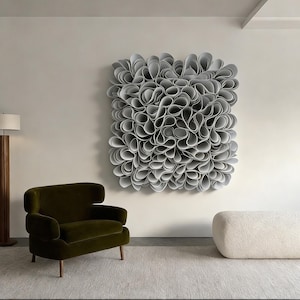 May include: A large, abstract wall sculpture made of numerous gray, petal-like forms. The artwork hangs on a white wall above a green armchair and a white, rounded ottoman. The room has a modern, minimalist design.