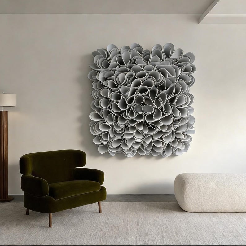 May include: A large, abstract wall sculpture made of numerous gray, petal-like forms. The artwork hangs on a white wall above a green armchair and a white, rounded ottoman. The room has a modern, minimalist design.