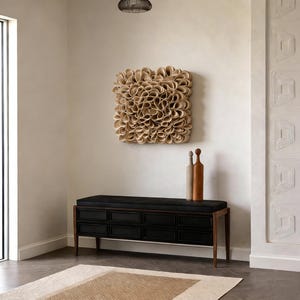 May include: A contemporary interior with a beige wall adorned with a square, textured artwork in beige tones. A black bench with drawers and wooden legs is below. Two decorative bottles are on the bench. A neutral rug and a window are also visible.