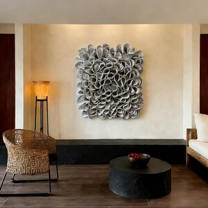 May include: A large, abstract wall art piece with a textured, layered design in shades of grey and white. The artwork is mounted on a beige wall, above a black bench. The room also features a woven chair, a black side table, and a floor lamp.