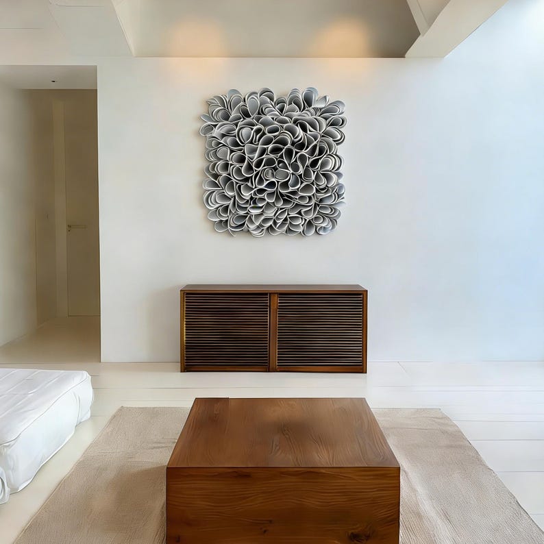 May include: A contemporary living space with a large, abstract wall sculpture composed of layered, petal-like forms in grey and white. Beneath, a dark wood cabinet with slatted doors is set against a white wall. A wooden coffee table and a beige rug complete the scene.