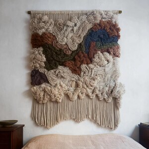 May include: A large, textured wall hanging with a neutral color palette. The artwork features various shades of beige, brown, and green, with a fringed bottom. The design incorporates a mix of woven and knotted textures, creating a three-dimensional effect. The wall hanging is mounted on a wooden rod.