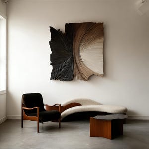 Op de afbeelding: Een moderne woonkamer met een groot, abstract kunstwerk aan de muur met een verloop van zwart, bruin en wit. Een donkerhouten fauteuil met een zwart kussen, een gebogen bank en een houten salontafel maken de scène compleet.