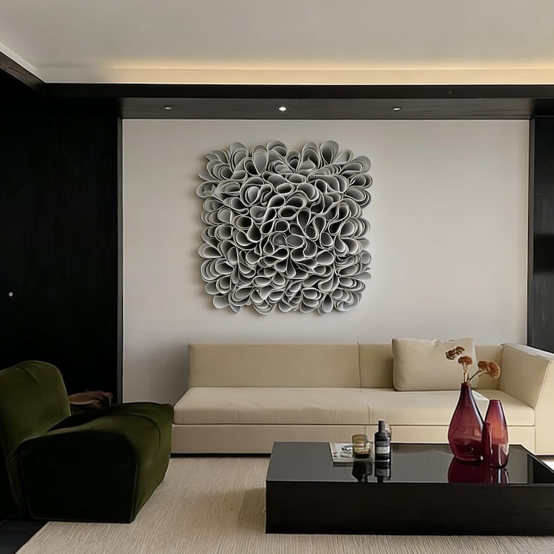 May include: A contemporary living room with a large, abstract wall sculpture made of layered, grey, petal-like forms. The room includes a beige sofa, a dark green armchair, and a black coffee table with decorative items.