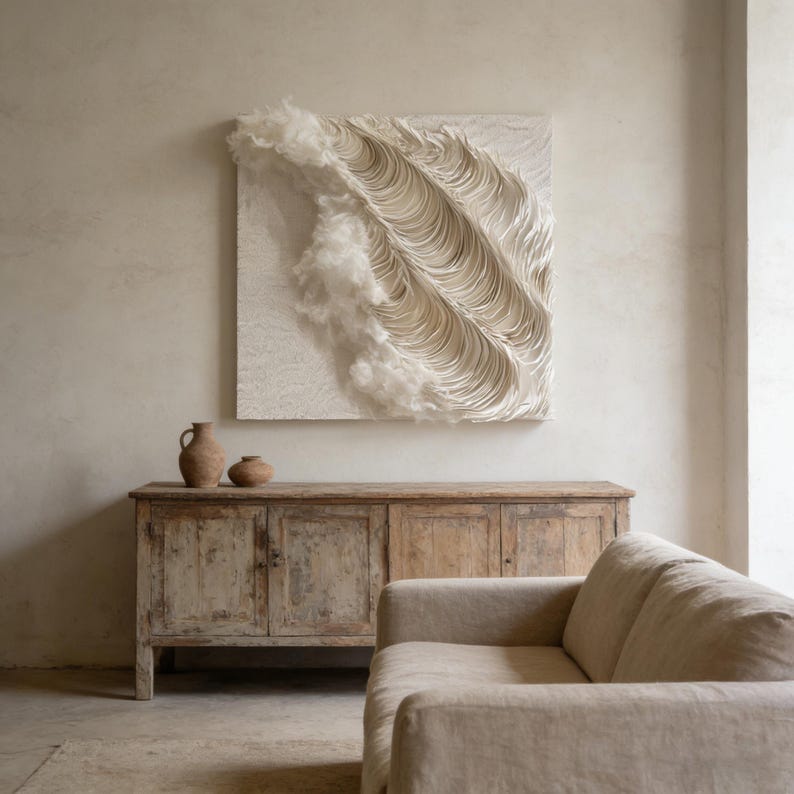 May include: A large, square, cream-coloured wall art piece with a textured, wave-like design. The artwork is mounted above a distressed wooden cabinet. Two small, brown ceramic vases sit on the cabinet. A beige sofa is in the foreground.