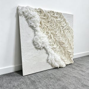 May include: A square, textured white canvas artwork with a three-dimensional design. The piece features a flowing, textured white element that cascades diagonally across the canvas, creating a visually dynamic and tactile experience. The artwork is mounted on a white wall.