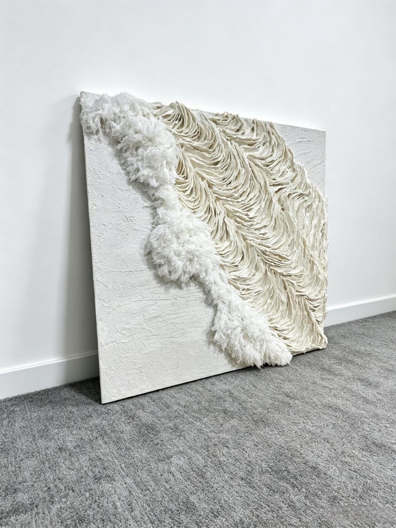 May include: A square, textured white canvas artwork with a three-dimensional design. The piece features a flowing, textured white element that cascades diagonally across the canvas, creating a visually dynamic and tactile experience. The artwork is mounted on a white wall.