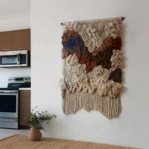 May include: A large, textured wall hanging with a landscape design. The artwork features a mix of beige, brown, green, and blue yarns, creating a three-dimensional effect. The bottom edge has a fringe.