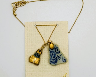 Pode incluir: Colar de corrente dourada com dois pingentes. Um pingente é uma peça de cerâmica de forma irregular de cor dourada. O outro pingente é uma peça de cerâmica branca com um desenho floral azul. A etiqueta anexada ao colar diz "tsugi et tsugu".