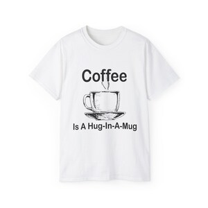 May include: White t-shirt featuring a black illustration of a coffee cup and saucer, with steam rising. The text "Coffee Is A Hug-In-A-Mug" is printed below the image. A casual, everyday wear item.