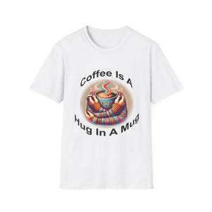 Peut inclure: T-shirt blanc avec un graphique d'une tasse à café avec des bras et des jambes qui se font un câlin. Le texte "Coffee is a Hug in a Mug" est imprimé sous la tasse.