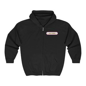 Könnte beinhalten: Schwarzer Kapuzenpullover mit Reißverschluss, Vordertasche und Kapuze. Der Hoodie hat einen kleinen Aufnäher auf der Brust mit dem Text "AIR FORCE" in weißen Buchstaben, umrahmt von einem rosa-goldenen Rand und Sternen.