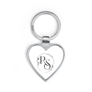 May include: Heart-shaped silver keychain with a circular design featuring the initials "RS" and the date "13-01-2026". The design includes floral accents. The keychain has a silver ring for attaching keys.
