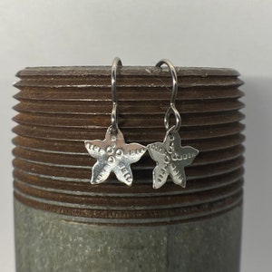 May include: A pair of silver starfish earrings with a textured surface. The earrings are hanging from a silver hook.