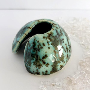 Puede incluir: Un jarrón de cerámica turquesa con una forma curva única. El jarrón tiene un patrón moteado de color marrón y verde y un acabado brillante. La abertura es un agujero oscuro y circular. Es un artículo decorativo.