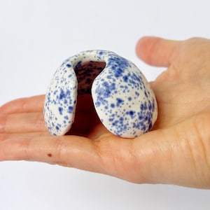 Könnte beinhalten: Kleine Keramikskulptur in Form eines gebogenen Bogens, gehalten in einer Hand. Die Skulptur ist weiß mit blauen Sprenkelmustern. Das Objekt ist ein dekoratives Element, möglicherweise für die Heimdekoration oder künstlerische Präsentation.