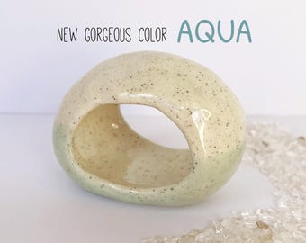 May include: A speckled, ceramic sculpture in the shape of a rounded donut. The sculpture is primarily cream-colored with black speckles and a light aqua-green gradient on the bottom. Text above reads "NEW GORGEOUS COLOR AQUA".