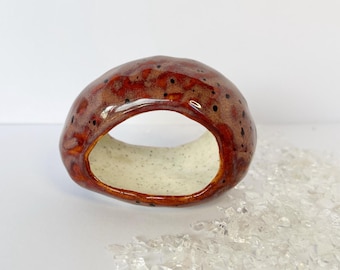 May include: A ceramic ring-shaped object with a glossy, speckled brown exterior and a cream-colored interior. The ring has a smooth, rounded form and is displayed on a white surface. Decorative accent.