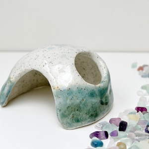 Puede incluir: Un escondite para mascotas de cerámica con un esmalte blanco moteado y verde azulado. La estructura arqueada tiene una abertura redondeada y una base de color verde azulado. Mide aproximadamente 13 cm de largo.
