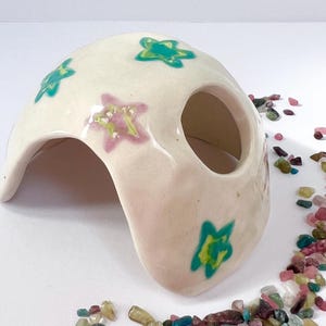 Könnte beinhalten: Eine gewölbte, höhlenartige Struktur aus cremefarbener Keramik mit einer kreisförmigen Öffnung. Die Struktur ist mit teal- und rosa-sternförmigen Designs verziert. Kleine, bunte Steine sind um die Basis der Struktur verstreut.