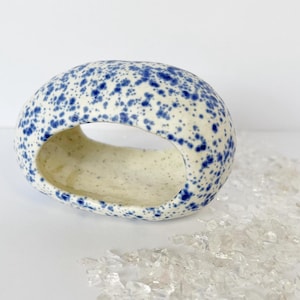 Puede incluir: Una pieza decorativa de cerámica, de forma ovalada, con interior y exterior color crema. El exterior está salpicado de puntos azules. La pieza tiene una gran abertura en el centro. Objetos transparentes, como cristales, están dispersos alrededor de la base.