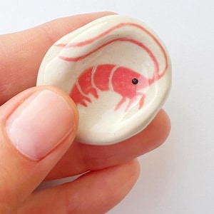 Puede incluir: Un pequeño plato de cerámica ovalado con un diseño de camarón pintado a mano. El camarón es rojo con un ojo negro, sobre un fondo blanco. El plato está sostenido en una mano.