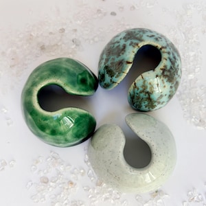 Puede incluir: Tres esculturas de cerámica en tonos verdes, turquesas y grises. Cada pieza tiene un diseño curvo y abierto. Las esculturas están dispuestas sobre una superficie blanca salpicada de pequeños cristales transparentes. El acabado brillante de la cerámica es visible.