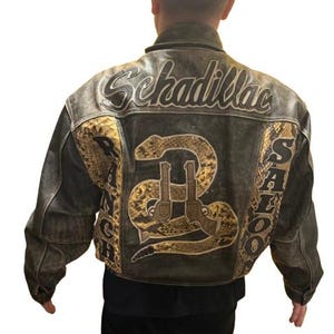 Könnte beinhalten: Eine braune Lederjacke mit einem Schlangen- und Pistolenmotiv auf der Rückseite. Die Jacke trägt die Worte "Schadillac Ranch Saloon" auf der Rückseite.