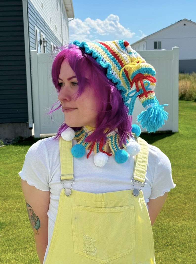 May include: A person wearing a colorful crocheted hat and matching neck scarf with pom-poms. The hat is striped with red, yellow, and blue, and the scarf has a similar pattern. The person is wearing a white shirt and yellow overalls.