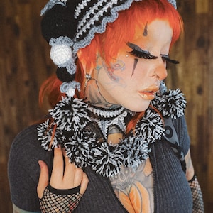 May include: A woman with red hair wearing a black and white crocheted hat with pom-poms. She is wearing a black shirt with a zipper and has tattoos on her neck, chest, and arms.