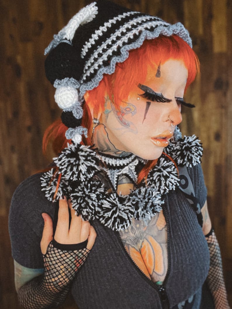 May include: A woman with red hair wearing a black and white crocheted hat with pom-poms. She is wearing a black shirt with a zipper and has tattoos on her neck, chest, and arms.