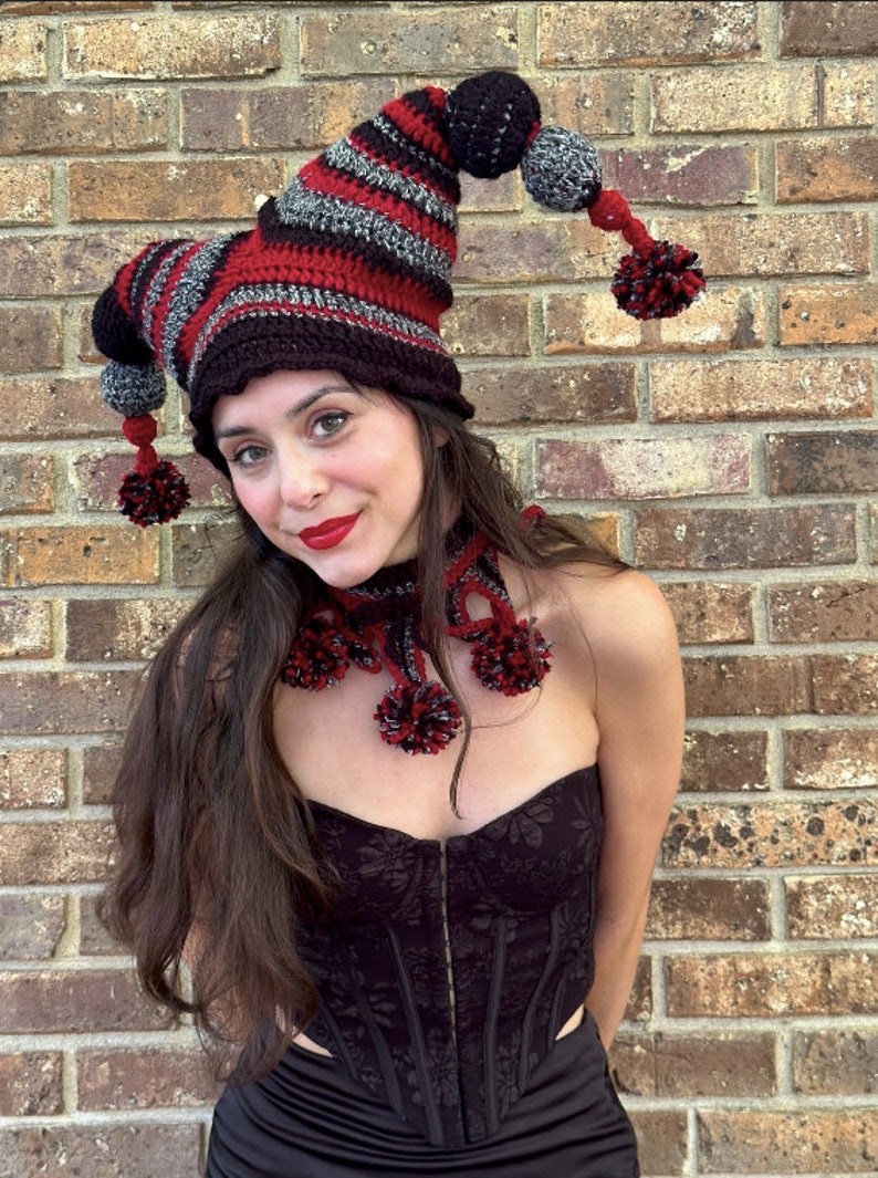 May include: A woman wearing a red and black striped crocheted jester hat with black and gray pom-poms. She is also wearing a black crocheted scarf with red pom-poms.