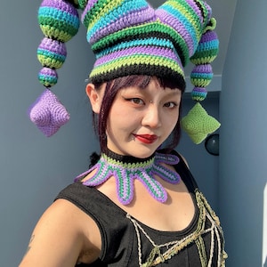 May include: A person wearing a colorful crocheted jester hat with a matching crocheted necklace. The hat has long, pointed, striped horns and the necklace has a pointed, scalloped design.