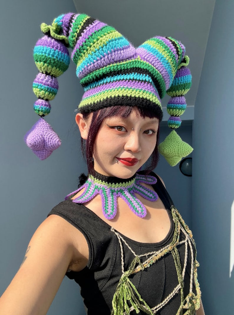 May include: A person wearing a colorful crocheted jester hat with a matching crocheted necklace. The hat has long, pointed, striped horns and the necklace has a pointed, scalloped design.
