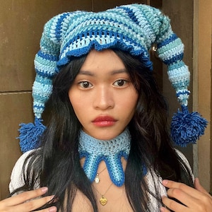 May include: A blue and white crocheted hat with a long, pointed tail and a pom-pom. The hat is worn by a person with long, dark hair and a blue crocheted neck warmer.