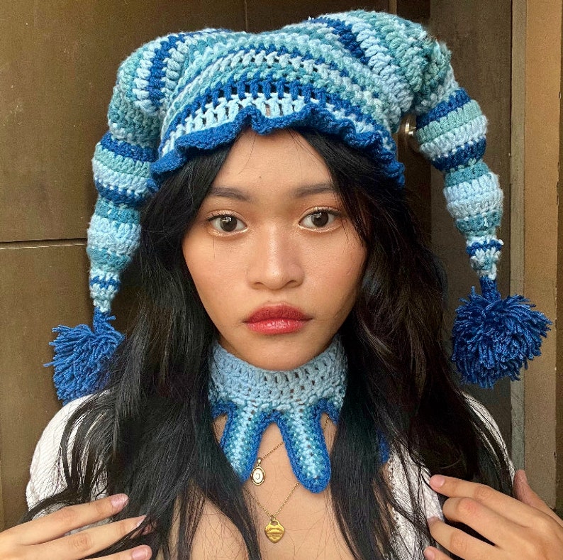 May include: A blue and white crocheted hat with a long, pointed tail and a pom-pom. The hat is worn by a person with long, dark hair and a blue crocheted neck warmer.