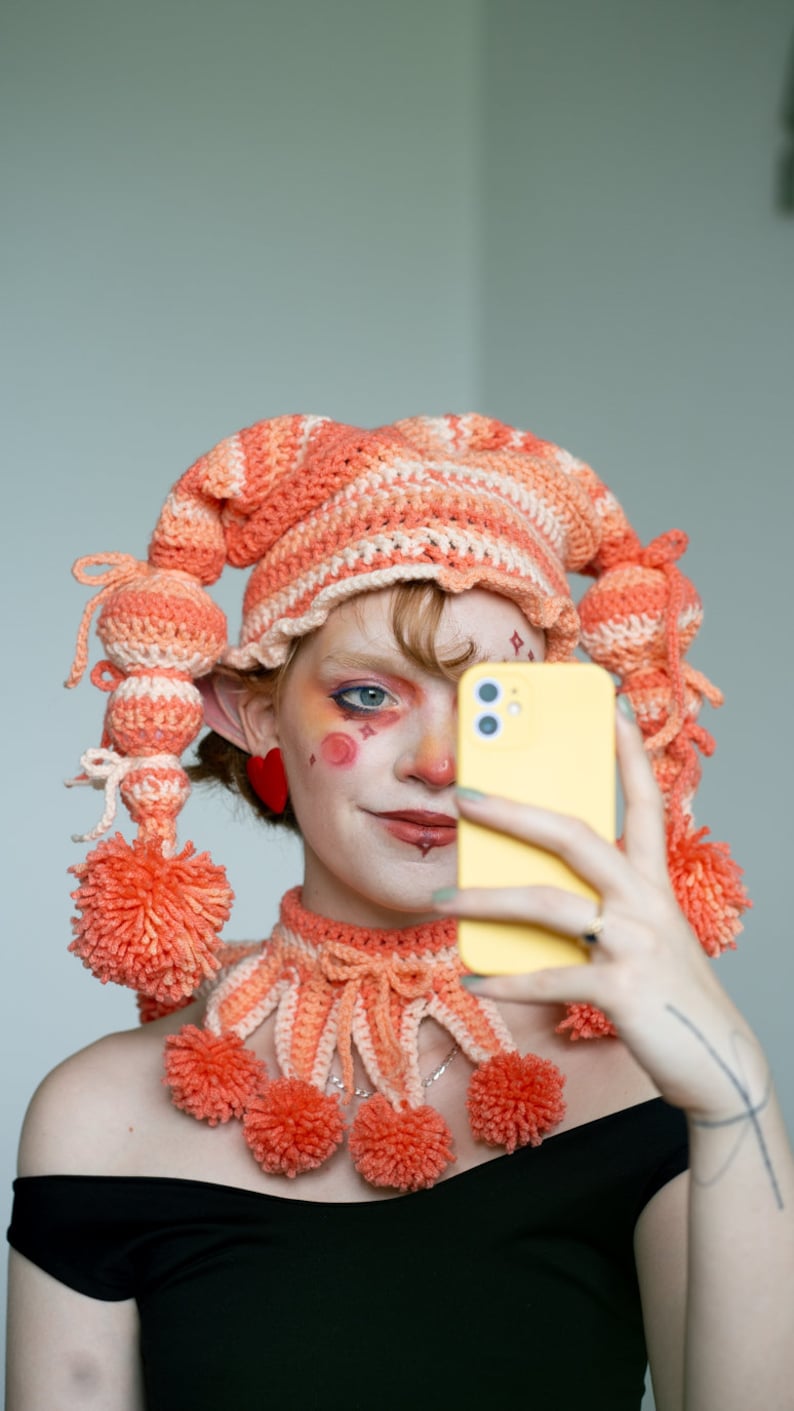 May include: A person wearing a crocheted hat and matching necklace with pom-poms in shades of pink and orange. They are holding a yellow smartphone in front of their face and smiling.