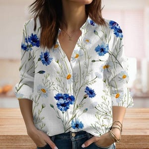May include: White button-down shirt with a floral design of blue cornflowers and white daisies. The long-sleeved shirt has a V-neck and is made of a lightweight fabric. The shirt is a casual style.