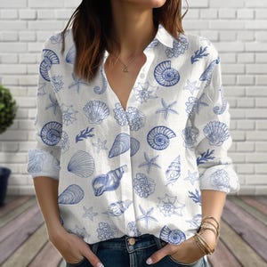 Puede incluir: Camisa blanca con estampado de conchas marinas azules. La camisa de manga larga presenta un cuello en V y un diseño repetido de varias conchas y estrellas de mar. La camisa está hecha de tela ligera.